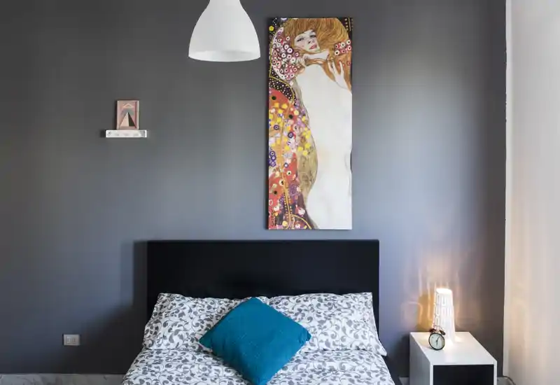 Camera da letto elegante con piumino a fantasia, cuscino accentuato turchese, testiera moderna e quadro decorativo. L'illuminazione del comodino e la composizione pulita creano un'atmosfera accogliente adatta a foto per annunci.