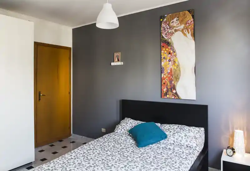 Camera da letto luminosa e ordinata con letto matrimoniale, cuscino decorativo e moderno quadro a parete. Presenti testiera scura, lampada da comodino e pavimento piastrellato pulito.
