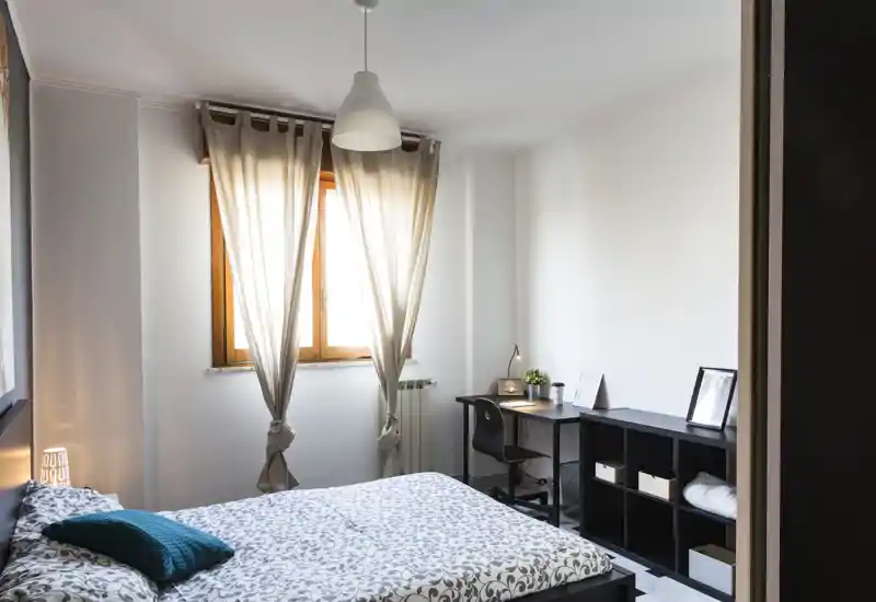 Camera da letto luminosa e curata con letto matrimoniale in primo piano, luce naturale dalla finestra con tende e una scrivania compatta con mensole lungo la parete.