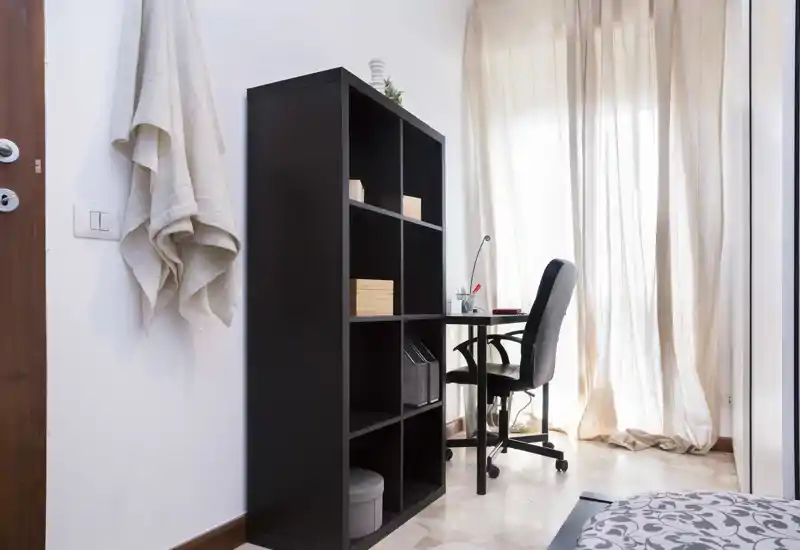 Angolo camera con zona studio: una libreria divide il letto da una scrivania compatta con sedia da ufficio davanti a tende chiare che lasciano passare la luce.
