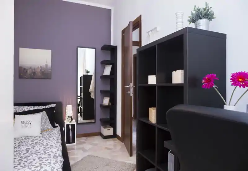 Camera da letto accogliente e ben arredata con letto singolo decorato, lampada da comodino, specchio a figura intera e mensole; parete accentuata viola e luce chiara e invitante.