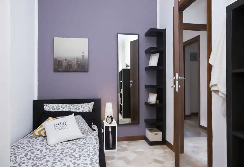 Camera singola luminosa e di stile con biancheria a motivi, cuscini decorativi e una lampada da comodino. Lo spazio include uno specchio alto, quadri e mensole che offrono spazio per riporre oggetti.