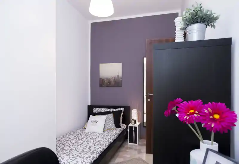 Camera singola luminosa e ben arredata con copriletto a fantasia, cuscini decorativi e lampada da comodino; armadio e fiori freschi rendono l'ambiente accogliente.