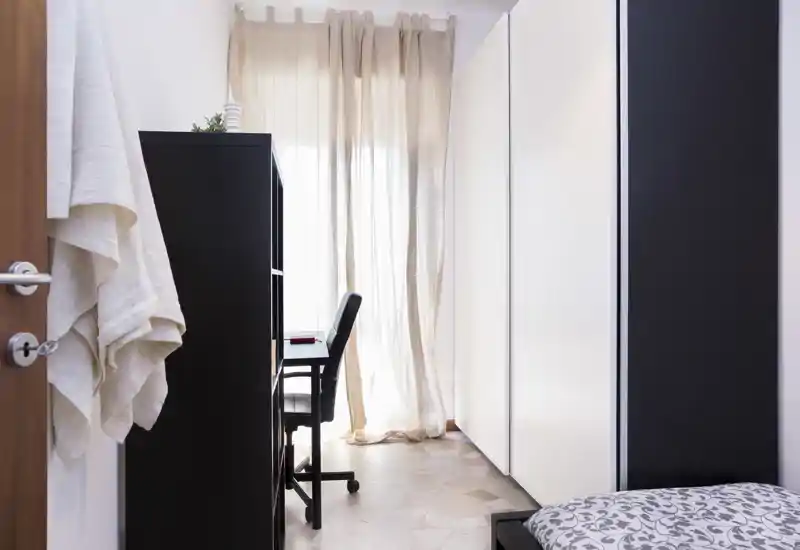 Angolo di camera da letto luminoso e compatto con letto singolo parzialmente visibile, scrivania e sedia vicino alla finestra con tenda leggera e armadi a parete. La stanza è ordinata e ben illuminata, adatta a un piccolo affitto o a studenti.
