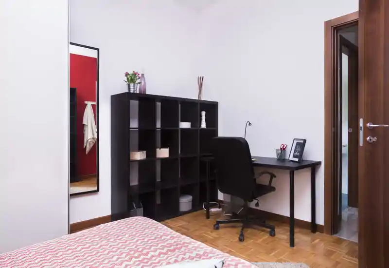Angolo camera luminoso e moderno con il letto in primo piano, una libreria nera e una scrivania compatta con sedia da ufficio — disposizione pratica ideale per uno studente o un occupante singolo.