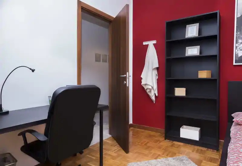Angolo della camera da letto con bordo del letto visibile, una libreria nera e una scrivania; parete rossa e parquet in legno per un aspetto moderno e accogliente.