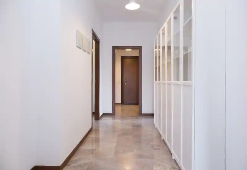 Ingresso luminoso e minimal con pavimento in pietra lucida, pareti bianche e armadi a muro su un lato — accesso pulito e ben illuminato alle stanze interne.