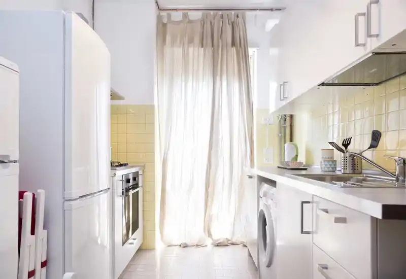 Cucina compatta e luminosa con mobili bianchi, rivestimento a piastrelle e tenda che filtra la luce naturale — dotata di piano cottura, frigorifero e lavello.