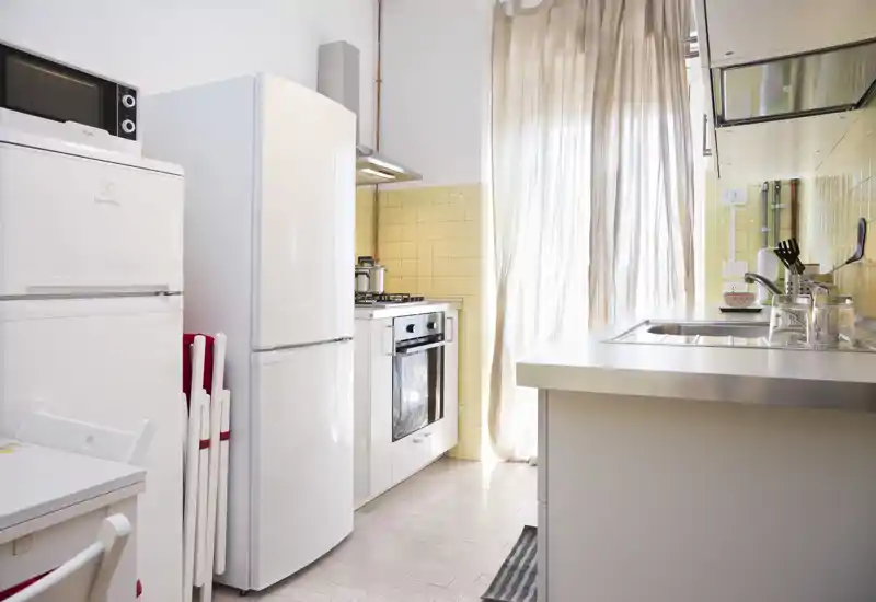 Cucina compatta e luminosa con elettrodomestici bianchi, forno a gas e lavello in acciaio. Tende leggere lasciano entrare luce naturale e piastrelle giallo chiaro donano calore.