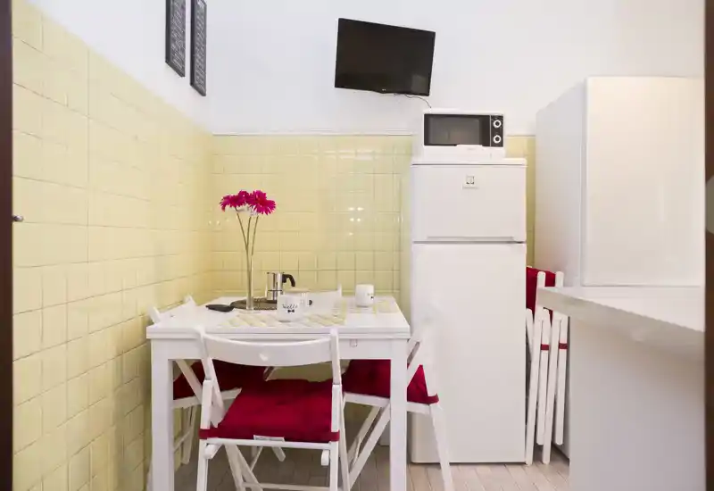 Angolo cottura compatto con un piccolo tavolo da pranzo bianco, sedie pieghevoli con cuscini rossi, frigorifero con microonde sopra, piastrelle giallo pallido e TV a parete — perfetto per un monolocale accogliente.