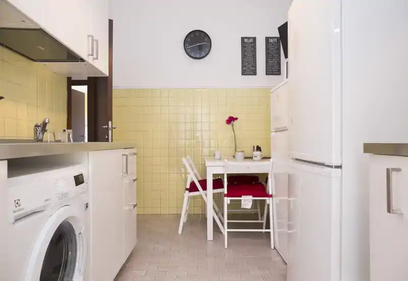 Cucina compatta e ordinata con paraschizzi in piastrelle giallo pallido, piccolo tavolo bianco con cuscini rossi, elettrodomestici integrati e lavatrice; ambiente luminoso e accogliente ideale per un appartamento.