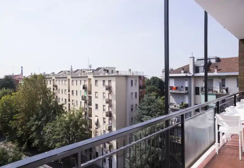 Balcone soleggiato con ringhiera in metallo e posti a sedere, con vista sugli edifici vicini e le chiome degli alberi — ideale per un caffè mattutino o per rilassarsi all'aperto.