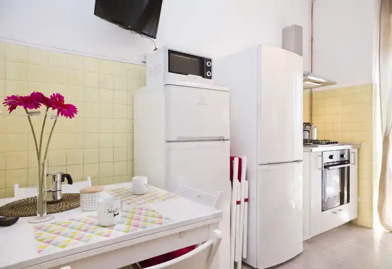 Cucina compatta e luminosa con elettrodomestici bianchi, piano cottura a gas e un piccolo tavolo da pranzo con tovagliette e fiori freschi — perfetta per pasti intimi.