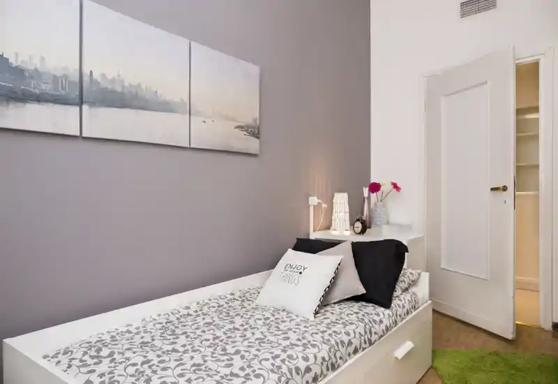 Dormitorio individual luminoso y bien decorado con cama hecha, cojines decorativos, mesita con lámpara y flores; tonos neutros y cuadro de pared que crean una atmósfera moderna y serena.