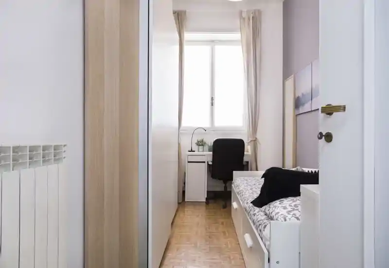 Habitación individual compacta y luminosa con cama tipo sofá, almacenamiento integrado y un pequeño escritorio junto a la ventana — ideal para estudiante o una sola persona.