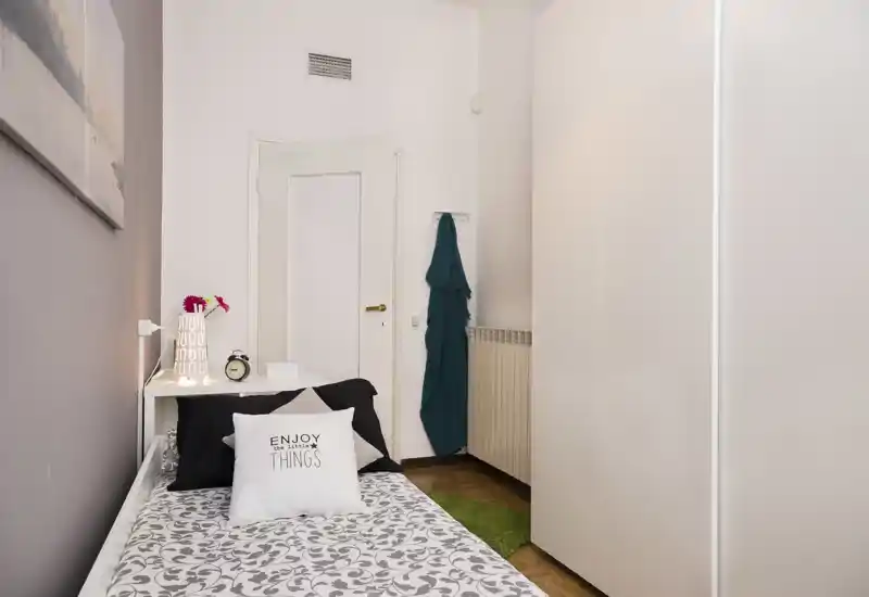 Habitación individual compacta y bien decorada con ropa de cama estampada, cojines decorativos y mesita con lámpara. Espacio luminoso y limpio, ideal para un alquiler acogedor.