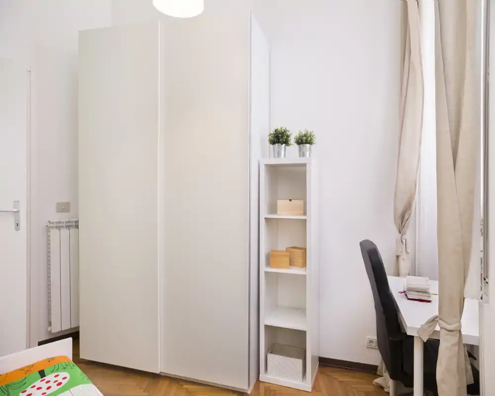 Angolo camera minimal e luminoso con un grande armadio bianco, una libreria stretta e una scrivania vicino alla finestra. Si vede solo un angolo del letto, l’ambiente è ordinato e ben illuminato.