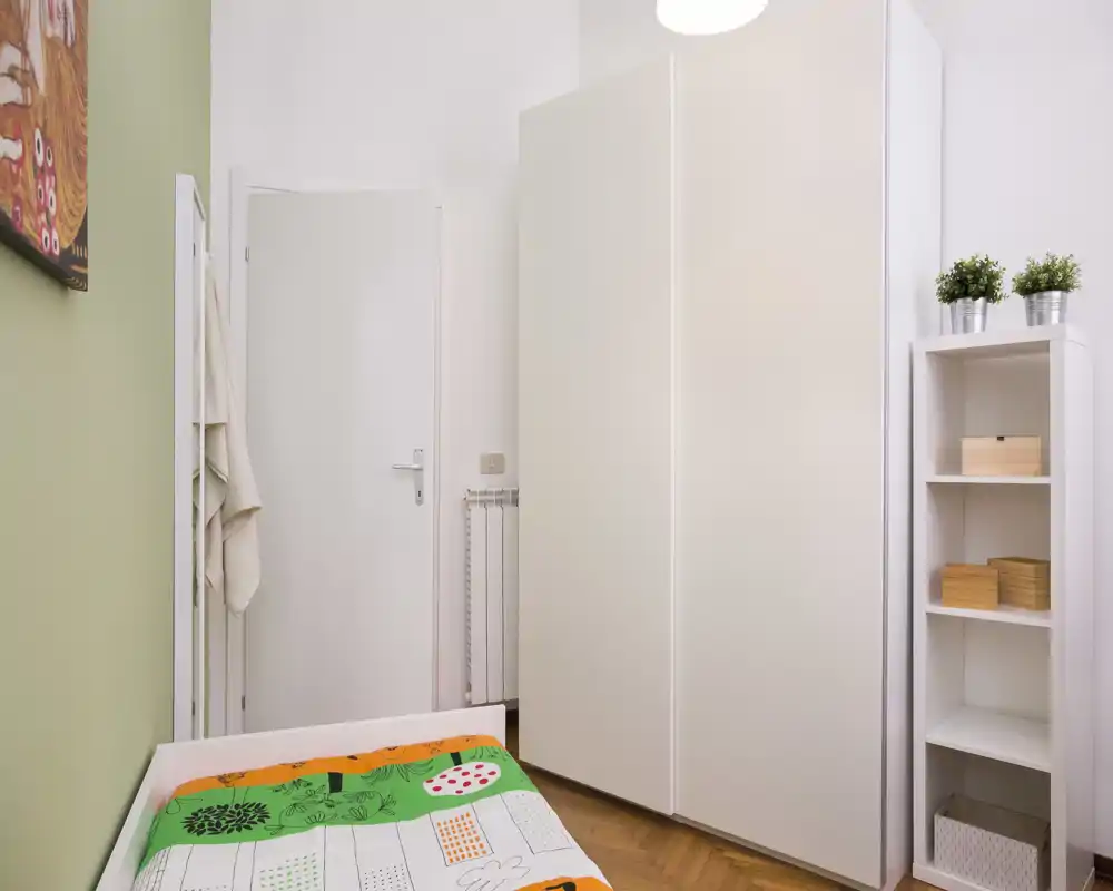 Piccola camera singola luminosa con una trapunta colorata, armadio bianco e scaffale; arredamento pulito e minimal ideale per un annuncio.