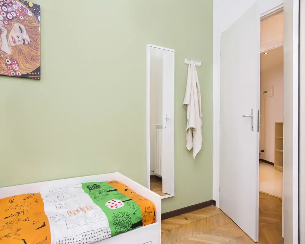 Piccola camera da letto con letto singolo contro una parete verde chiaro, biancheria colorata a fantasia, uno specchio a figura intera e una porta aperta verso un corridoio luminoso. Pulita, essenziale e ben illuminata — adatta per uno studente o una persona sola.