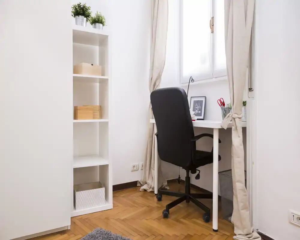 Angolo ufficio luminoso e compatto con scrivania bianca, sedia nera ergonomica, scaffale e luce naturale dalla finestra — perfetto per smart working o studio.