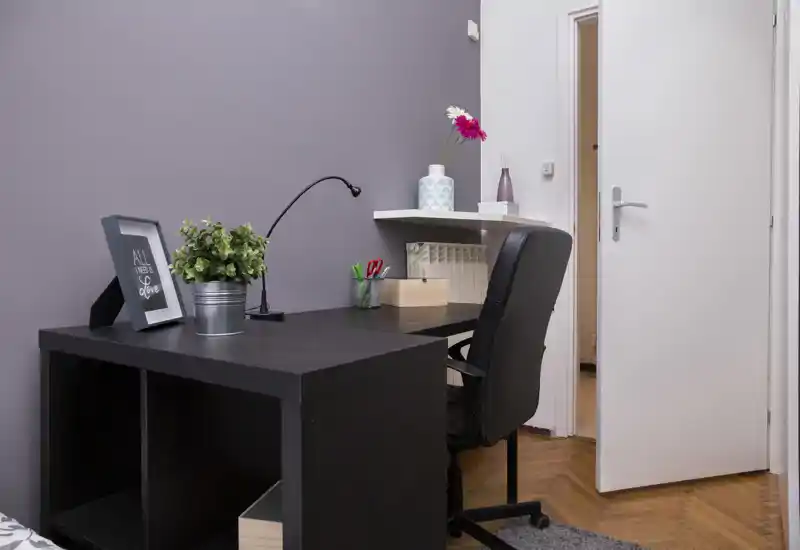 Angolo camera con scrivania in legno scuro, sedia girevole e semplici elementi decorativi (pianta, cornice, lampada). La porta aperta e il pavimento in parquet suggeriscono la disposizione della stanza.