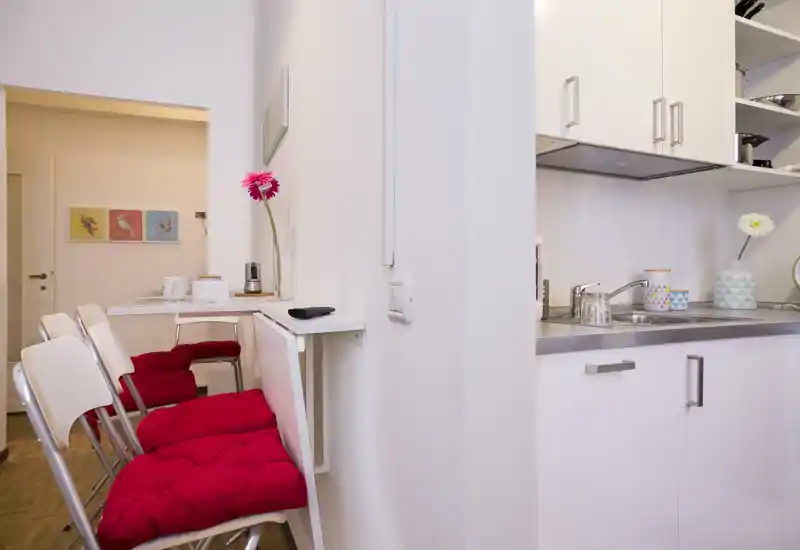 Angolo cottura compatto e luminoso con mobili bianchi, lavello in acciaio e un banco colazione con sedie pieghevoli con cuscini rossi — ideale per un piccolo appartamento.