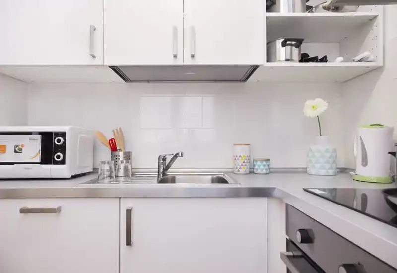 Cucina moderna e compatta con lavello in acciaio, microonde, piano a induzione e contenitori decorativi; superfici luminose e pulite con piccoli accenti come un vaso che rendono l'ambiente accogliente.