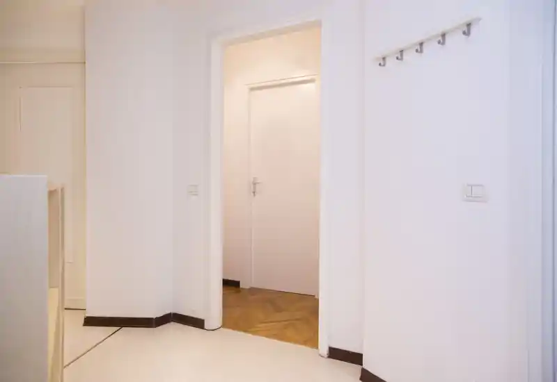 Ingresso luminoso e minimale con pareti bianche, parquet visibile attraverso la porta aperta e appendiabiti a parete.