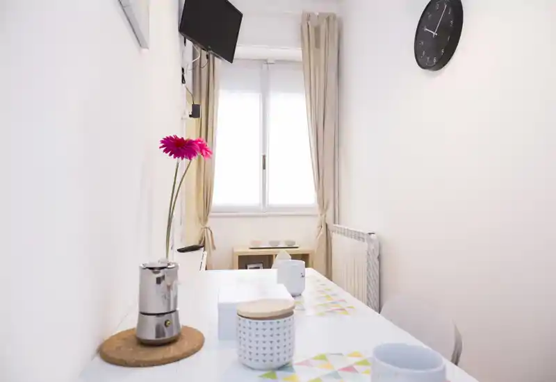 Piccola zona pranzo luminosa con un tavolo stretto preparato per un uso informale — tazze decorative, tovagliette e un fiore rosa creano un'atmosfera fresca e ordinata vicino alla finestra.