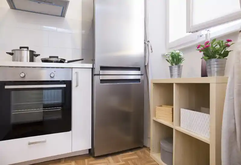 Cucina moderna compatta con elettrodomestici in acciaio inox, forno e frigorifero integrati, mensola in legno e piante vicino a una finestra luminosa.