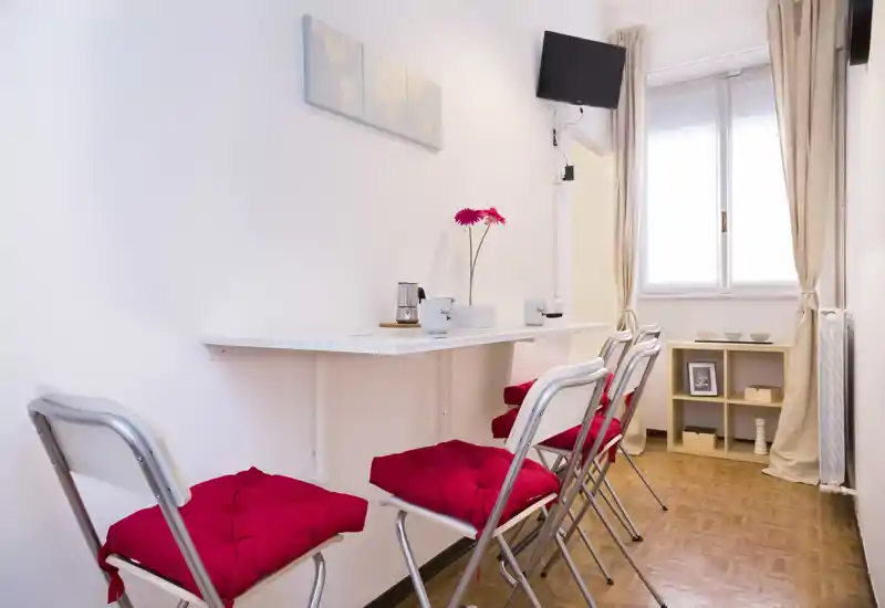 Zona pranzo compatta e luminosa con tavolo alto a parete, quattro sedie pieghevoli con cuscini rossi e una finestra che fornisce luce naturale. Arredamento semplice e moderno con un piccolo scaffale e dettagli decorativi.