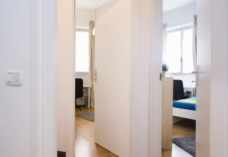 Camera da letto luminosa e compatta vista attraverso una porta aperta: letto singolo visibile, scrivania e finestra con luce naturale — ambiente ordinato e funzionale per una persona.