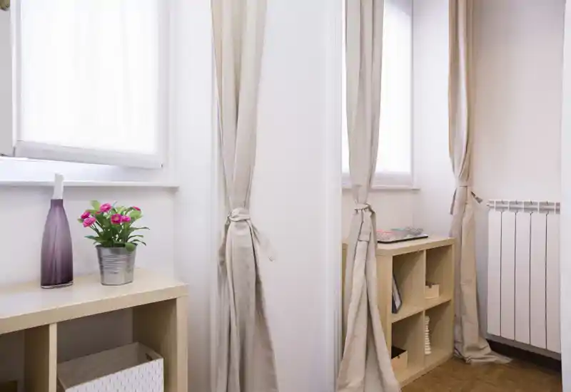 Angolo soggiorno luminoso e minimal con tende chiare, scaffalature in legno e dettagli decorativi come un vaso e piccoli fiori in vaso che creano un'atmosfera calma e ariosa.