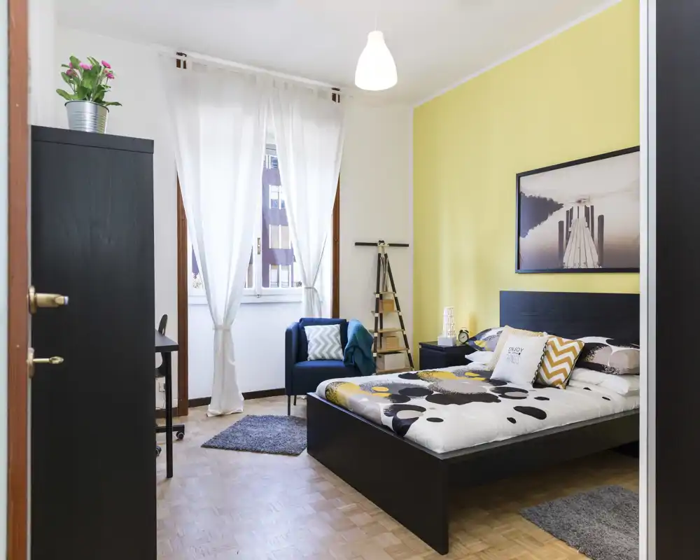 Dormitorio moderno y luminoso con cama doble, ropa de cama estampada y cojines coordinados; luz natural desde la ventana, un sillón acogedor y mobiliario negro minimalista.