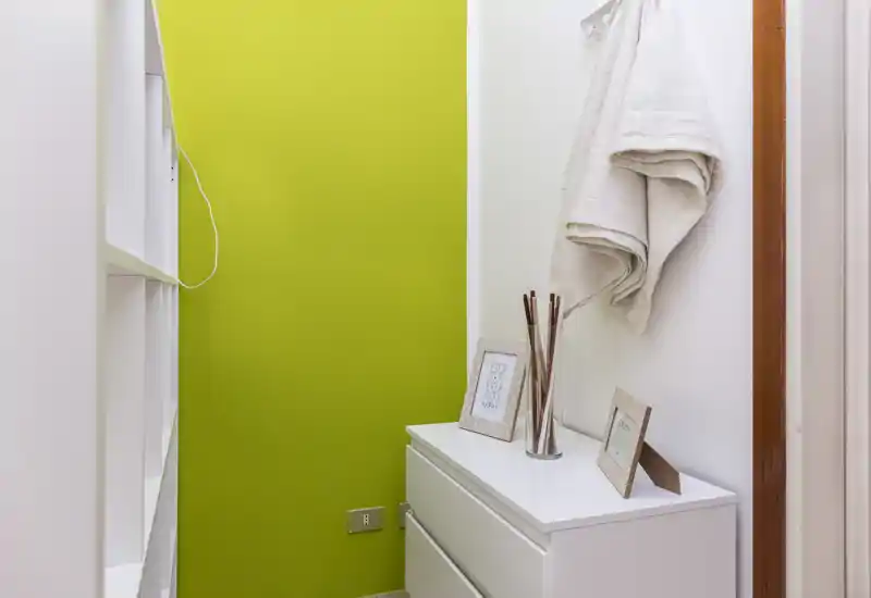 Pequeña entrada/pasillo ordenado con una cómoda blanca, marcos decorativos y textiles colgados frente a una pared verde llamativa.