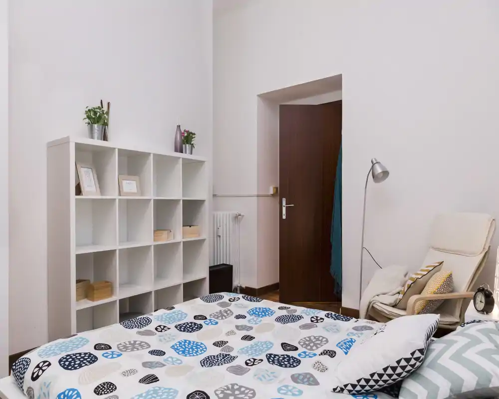 Bright, neatly styled bedroom with a patterned bedspread, shelving unit and a comfortable armchair. The image highlights the bed and a calm, welcoming atmosphere suitable for rental listings.