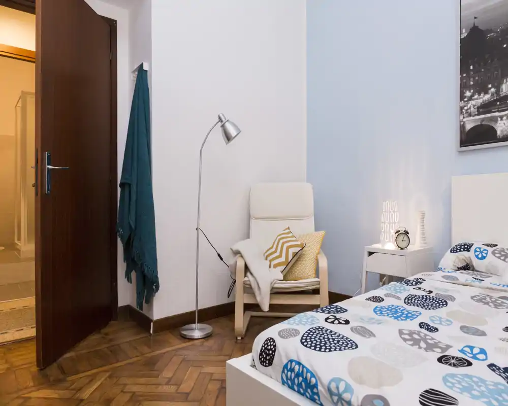 Well-staged bedroom corner with a patterned duvet, bedside table and a reading chair with cushions and lamp — cozy and bright.