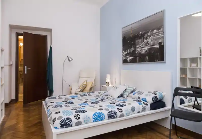 Camera da letto luminosa e curata con letto bianco, biancheria a fantasia e una poltrona accogliente; illuminazione soffusa e arredi semplici creano un ambiente invitante.