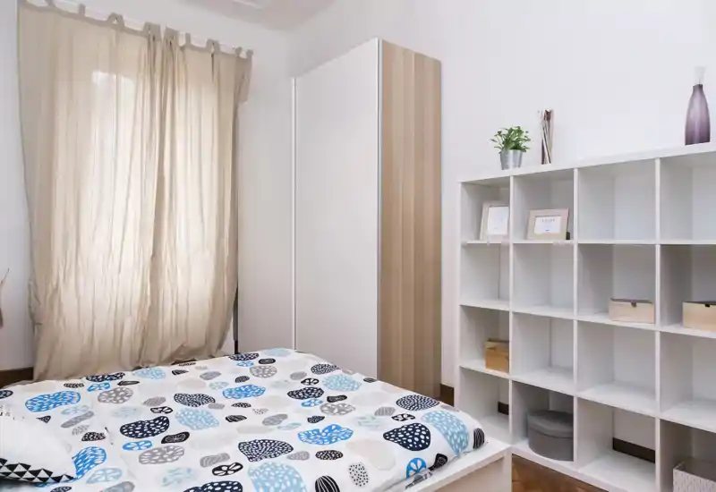 Camera da letto moderna e luminosa con copriletto a fantasia, armadio alto e scaffalatura a cubi; la luce naturale filtrata dalle tende crea un’atmosfera calma e accogliente.