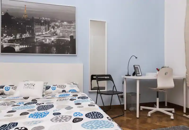 Camera da letto luminosa e ben arredata con copriletto a fantasia, cuscino decorativo e una piccola scrivania — ideale per soggiorni brevi o studenti.