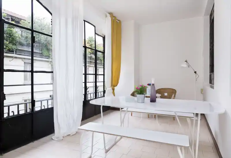 Área de comedor moderna y luminosa con una larga mesa blanca, banco y sillas de mimbre junto a grandes ventanales de estilo industrial que inundan el espacio de luz natural.