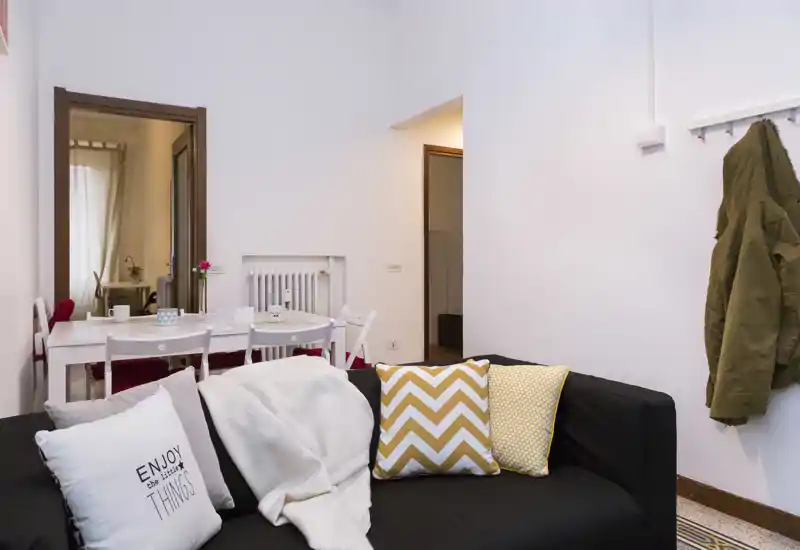 Sala luminosa y ordenada con un sofá negro decorado con cojines estampados y una manta; al fondo se aprecia una pequeña mesa de comedor que aporta una sensación acogedora.