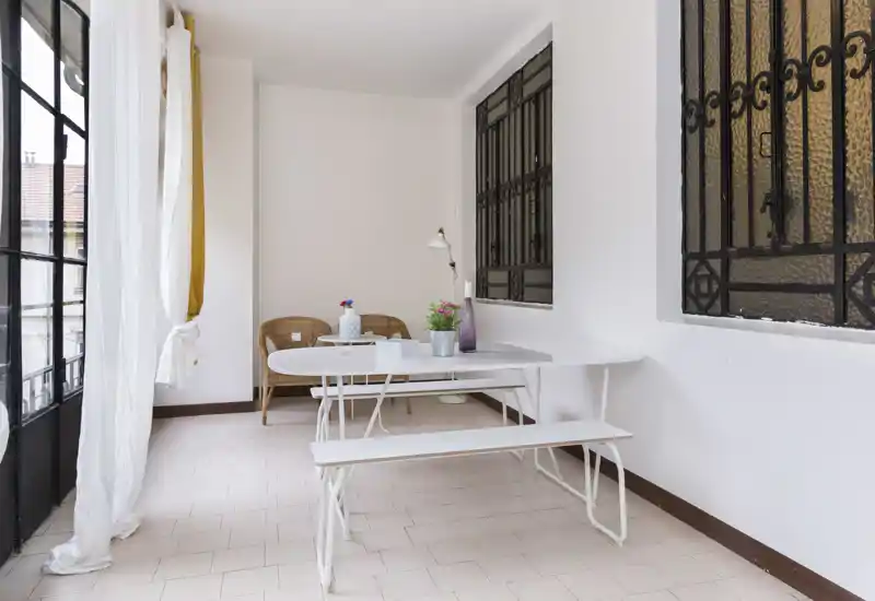 Zona de comedor luminosa y minimalista con mesa blanca y banco, sillas de mimbre y grandes ventanas que aportan luz natural — espacio sencillo y aireado, ideal para comidas informales.