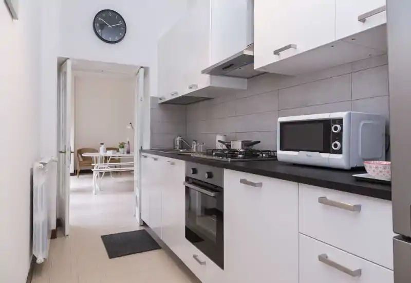 Cocina moderna tipo galería con armarios blancos, encimera negra, placa de gas y microondas; luminosa y ordenada con vista al comedor.