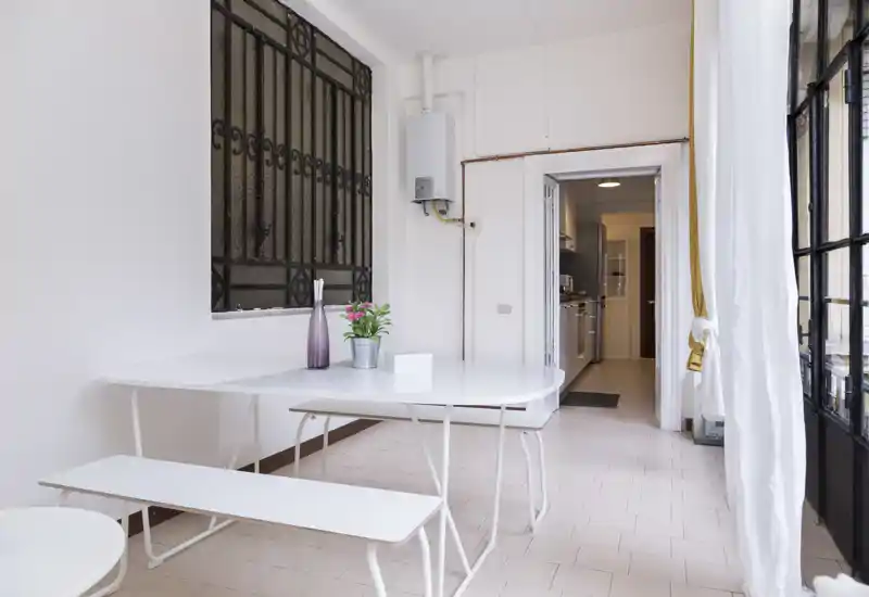 Zona de comedor minimalista y luminosa con mesa y banco blancos, jarrón decorativo y planta junto a una gran ventana/puerta que aporta luz natural.