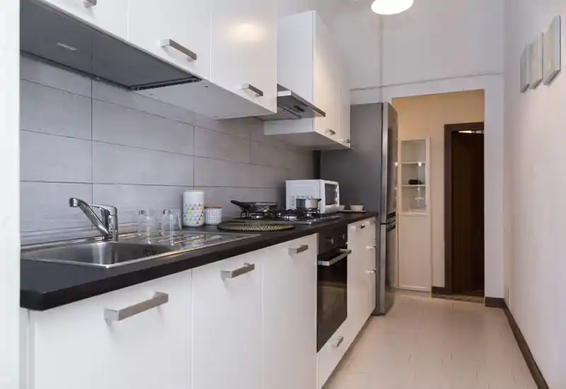 Cocina moderna tipo pasillo con muebles blancos, encimera negra, horno empotrado y frigorífico al fondo. Espacio limpio y ordenado con pequeños tarros decorativos y utensilios sobre la encimera.