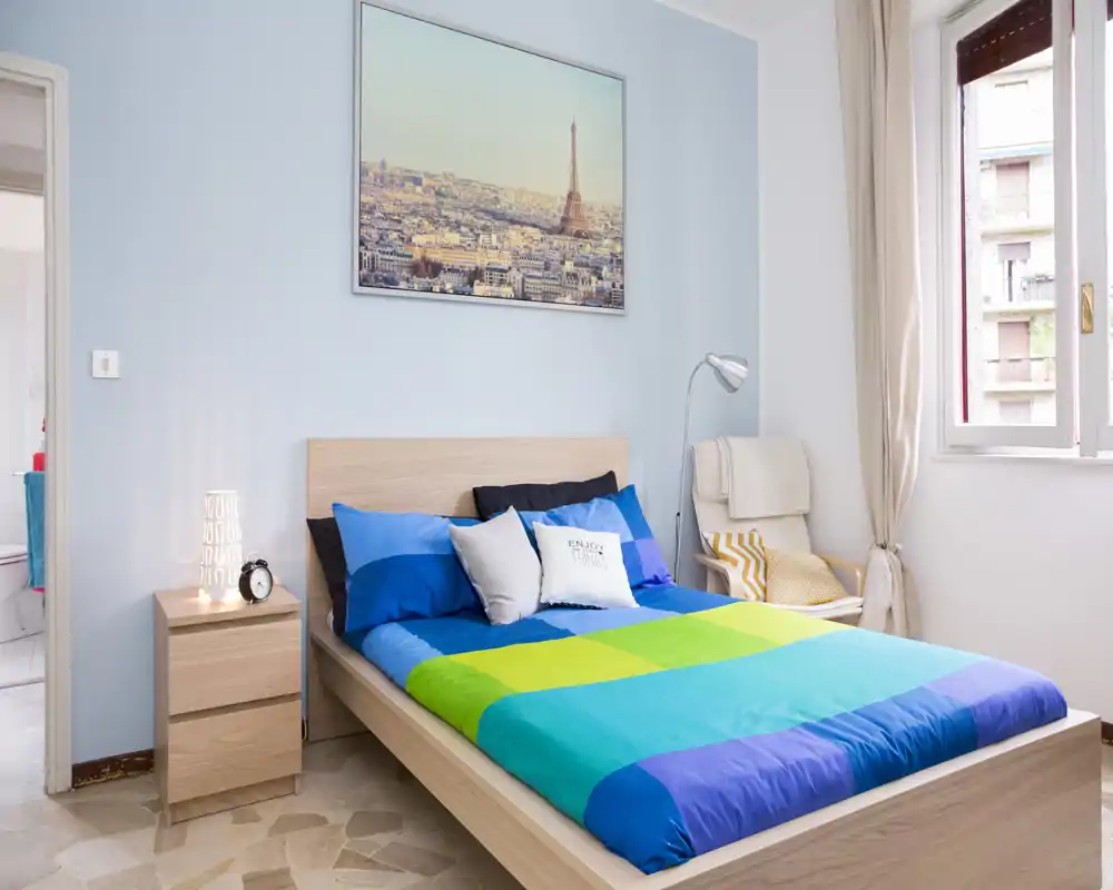 Bright, tidy bedroom with a wooden double bed, colorful bedspread, bedside table with lamp and a reading chair by the window.