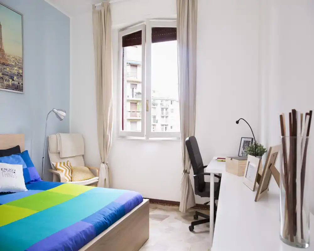 Bright, modern bedroom with a colorful double bed, reading chair and a work desk by the window — ideal for a comfortable stay with natural light.