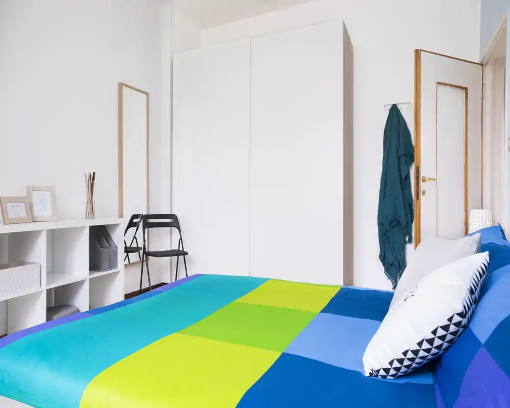 Bright, neatly staged bedroom with a colorful bedspread, large wardrobe and minimal modern furniture — ideal for rental photos.