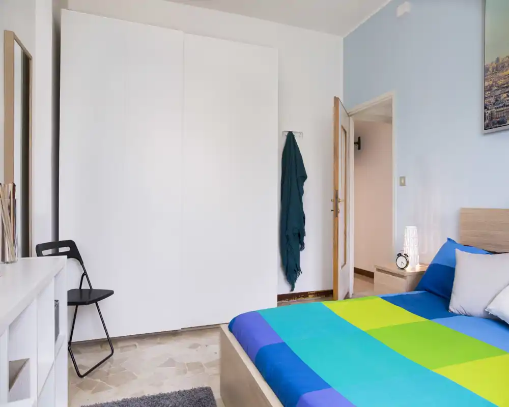 Bright, neatly staged bedroom with a colorful double bed, bedside table and wardrobe — ideal for rental listings emphasizing fresh décor and natural light.